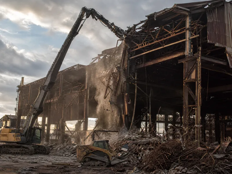 A high-reach excavator with a shear attachment tearing down a steel mill building